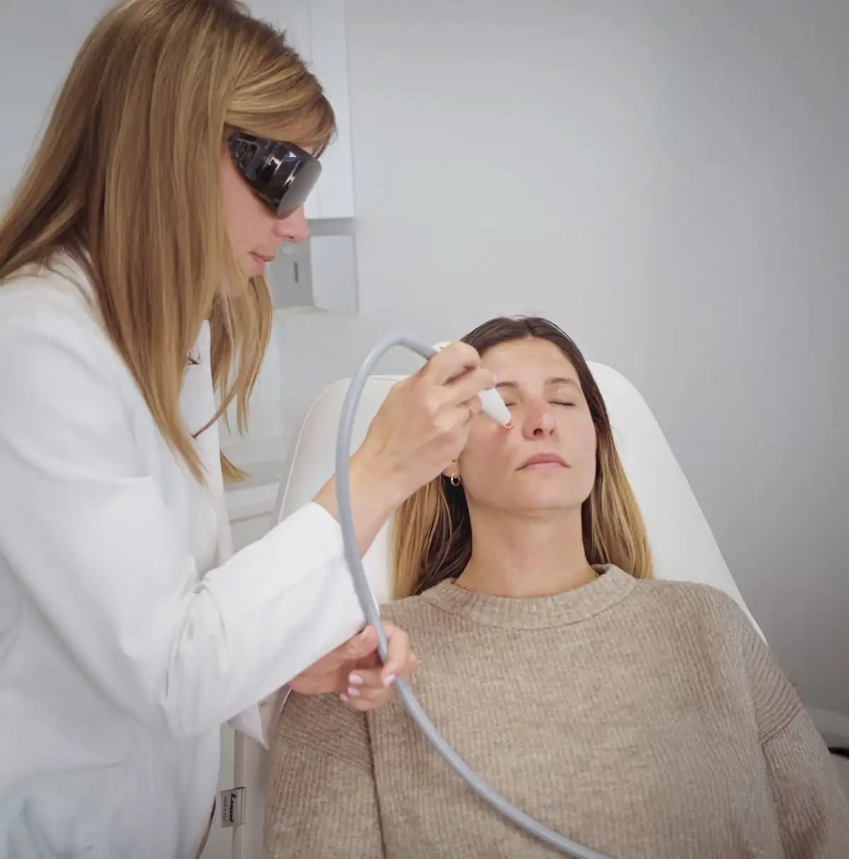 Le Docteur Bela en blouse blanche et lunettes de protection effectue un traitement de lumière intense pulsée à l’aide d’un appareil à main sur une patiente allongée, les yeux fermés, dans un cabinet médical
